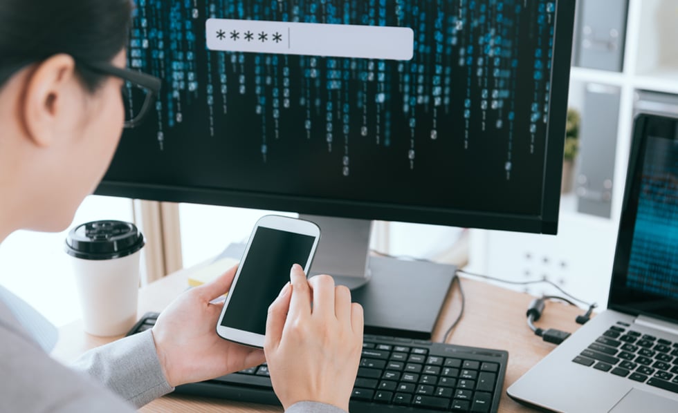 Man sits in front of a computer screen filled with code while looking at his iphone