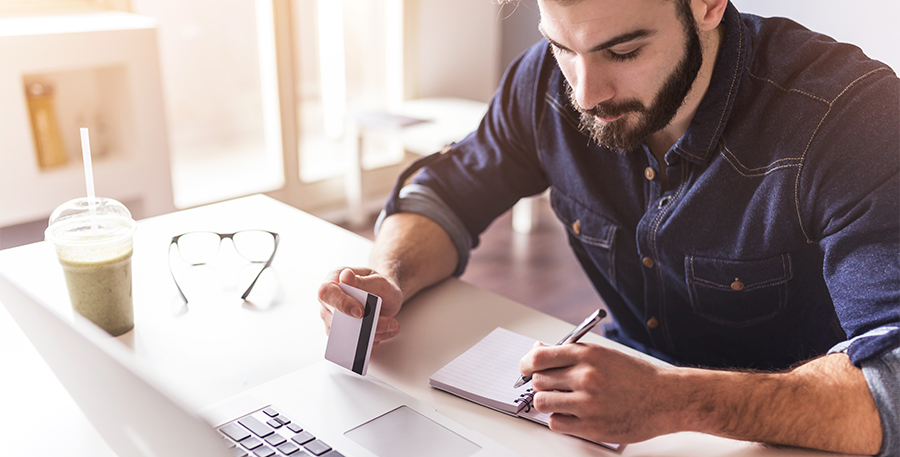 Man in a denim short sitting at a desk while jotting down notes in his note pad with a credit card in the other hand