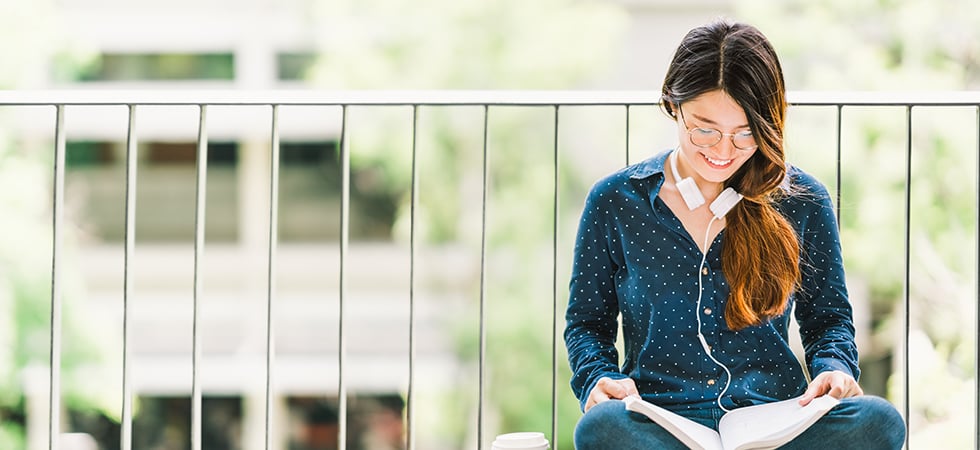 Woman with ling hair in a blue shirt and glasses reading a book on a balcony outside